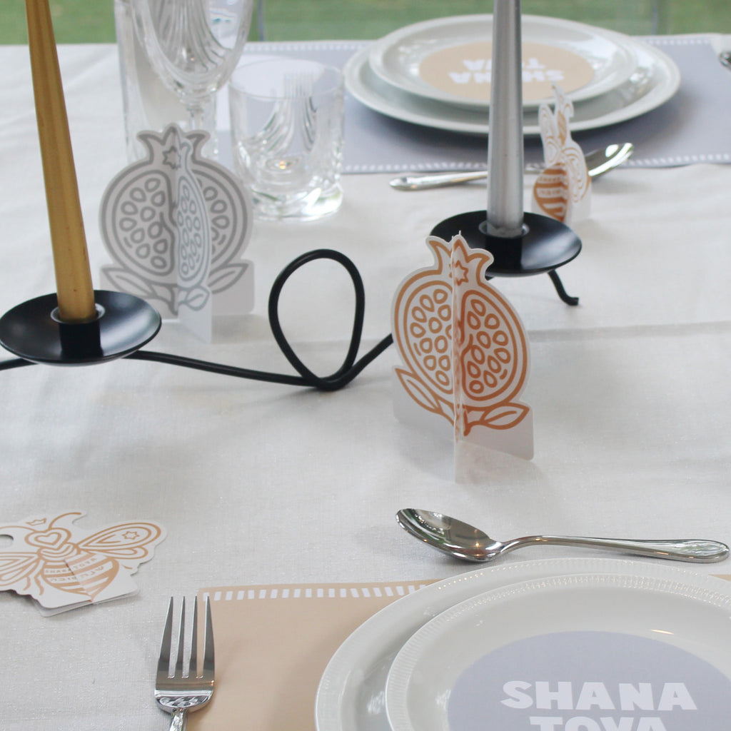 A styled table setting with white plates, silver cutlery, clear glassware, and decorative paper pomegranate place cards on a white tablecloth. A round plate in the foreground reads 'SHANA TOVA'.