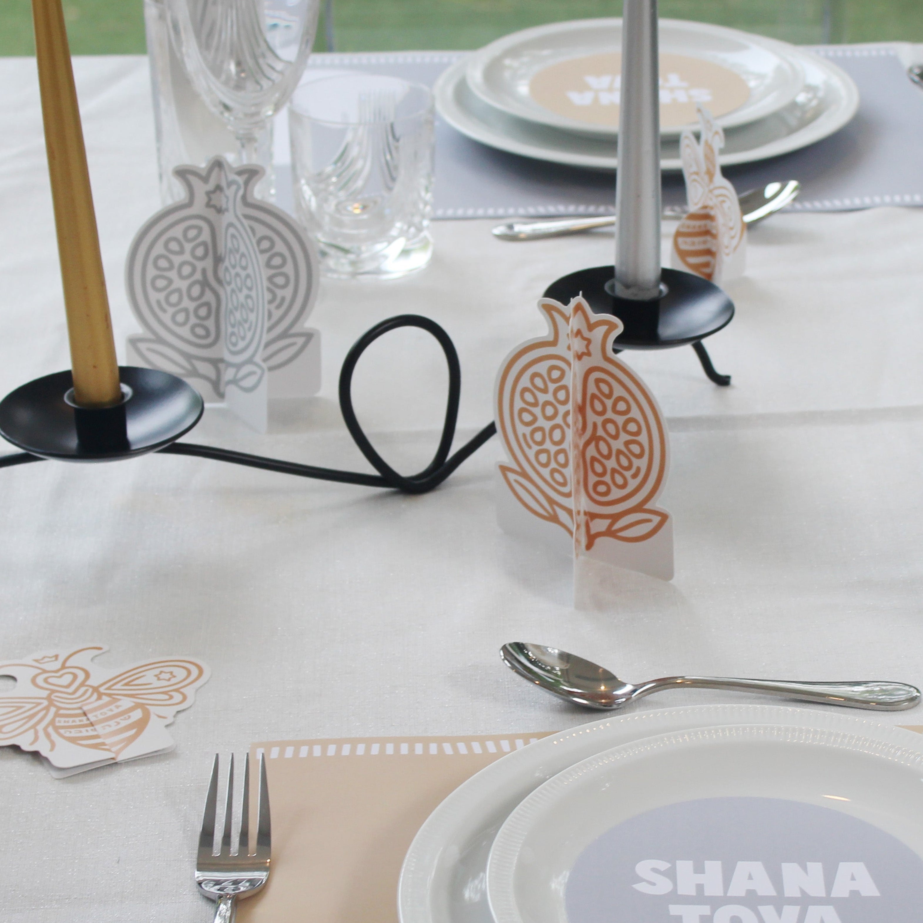 A styled table setting with white plates, silver cutlery, clear glassware, and decorative paper pomegranate place cards on a white tablecloth. A round plate in the foreground reads 'SHANA TOVA'.
